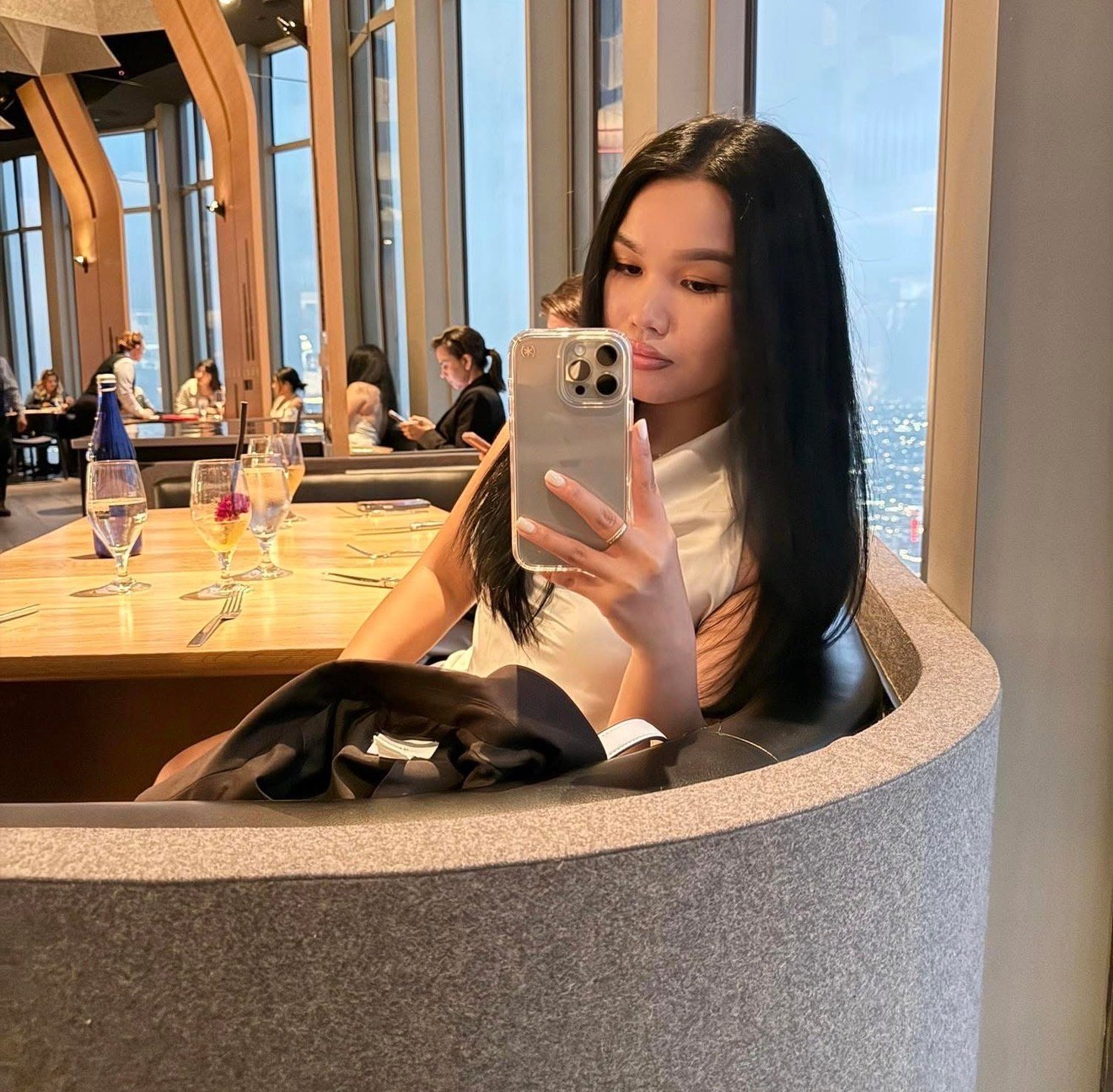 Woman with long black hair taking a selfie in a modern high-rise restaurant with floor-to-ceiling windows and city views